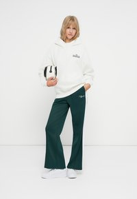 Weißer Fleece-Hoodie mit grünem Logo, kombiniert mit dunkelgrünen, weitem Beinen und weißen Seitenstreifen sowie weißen Sneakers. Kleine schwarz-weiße Tasche.