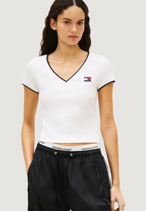 Kobieta ubrana w biały krótki t-shirt z dekoltem w kształcie litery V z czarną lamówką oraz czarne spodnie z troczkami, prezentująca markowany pas z napisem „tommy jeans”.