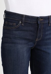 Tmavě modré džíny z denimu mají klasický rovný střih, viditelné švy, zapínání na knoflík vpředu a dvě boční kapsy.