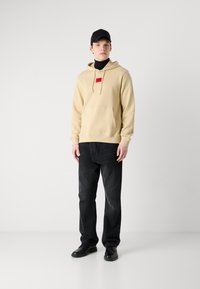 Sudadera beige con un bolsillo frontal y un logotipo rectangular rojo, combinada con jeans negros y zapatillas negras, contra un fondo neutro.
