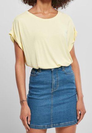 Femme portant un chemisier jaune pâle aux manches retroussées, rentré dans une mini-jupe en jean bleu taille haute, debout devant un fond neutre.