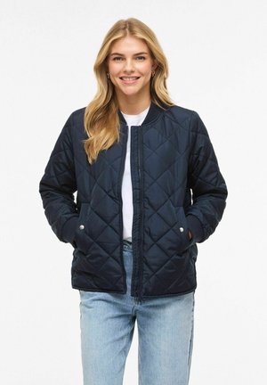 Jeune femme aux longs cheveux blonds portant une veste matelassée bleu marine, une chemise blanche et un jean bleu clair, souriante, les mains dans les poches sur fond blanc.