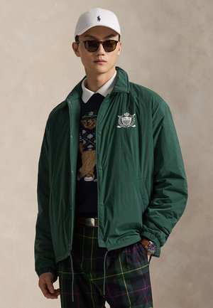 Mand iført grøn jakke med emblem, ternede bukser, bjørnesweater, hvid kasket, solbriller og ur, stående med den ene hånd i lommen.