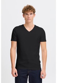 Tânăr cu păr creț, îmbrăcat într-un tricou negru simplu cu gât în V și blugi dark blue, stând în fața unui fundal alb.