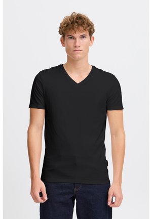 Ungdommelig mann med krøllete hår, iført en enkel svart V-hals kortermet T-skjorte og mørkeblå jeans, stående mot en hvit bakgrunn.