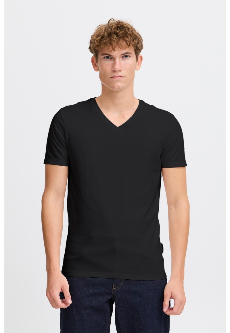 Giovane uomo dai capelli ricci che indossa una semplice maglietta nera a V con maniche corte e jeans blu scuro, in piedi contro uno sfondo bianco.