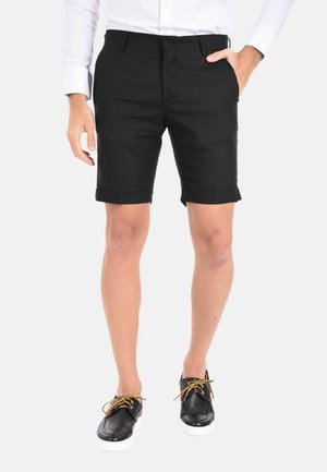 Uomo che indossa pantaloncini neri su misura, camicia bianca a maniche lunghe e scarpe da barca nere con suole bianche e lacci beige.