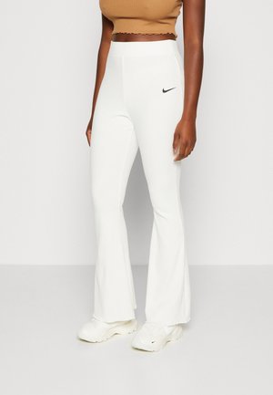 Žena v bílých vysokým pasem zdobených rozšířených kalhotách s černým logem Nike, béžovém krátkém topu a bílých teniskách stojící na bílém pozadí.