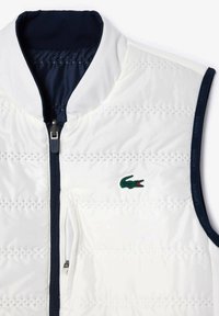 Chaleco de plumas blanco con un cuello azul marino, patrón de acolchado horizontal, un bolsillo con cremallera lateral y un logo de cocodrilo verde en el lado izquierdo del pecho.