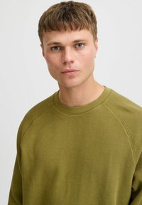 Hanorac tricotat de culoare verde măsliniu, cu țesătură texturată, guler rotund și mâneci raglan, având un design ajustat și o ușoară reverb.