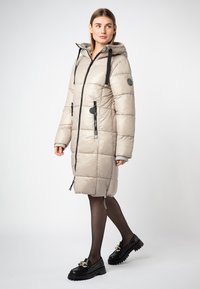 Lichtbeige pufferjas met capuchon, voorzien van zwarte trekkoorden, ritsen en mouwen tot de elleboog. Gequilte ontwerp en een knielange snit.
