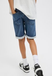 Shorts di denim blu scuro con orlo risvoltato, dotati di due tasche posteriori e cuciture a contrasto, abbinati a calzini bianchi e sneakers nere.