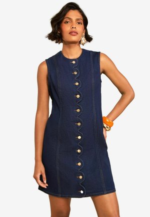 Woman wearing a sleeveless dark blue denim dress with scalloped button closure down the front and orange and gold bracelets on her wrist.
