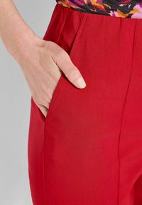 Hand mit natürlichen Nägeln, die in der vorderen Tasche einer leuchtend roten Hose ruht, getragen mit einem bunten Blumenshirt.