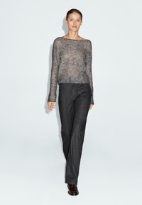 Donna che cammina in avanti indossando un ampio maglione grigio a maglia, pantaloni dritti grigio scuro e scarpe in pelle scura su uno sfondo bianco.