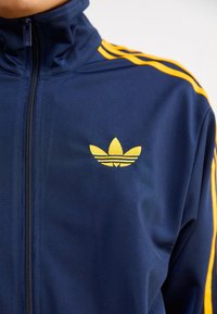 Námořnická modrá sportovní zipová bunda s žlutými třemi pruhy a logem, vyrobená z hladkého, lehkého materiálu. Vysoký límec.