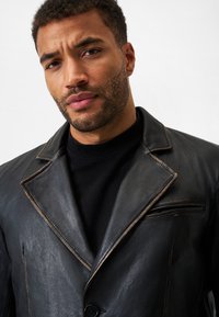 Schwarze Lederjacke mit strukturiertem Kragen, kontrastierenden braunen Nähten und einer Brusttasche. Der Stoff hat eine glatte, strukturierte Oberfläche.