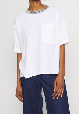 Personne portant un t-shirt blanc oversize avec une poche poitrine et un col rayé arc-en-ciel, associé à un jean bleu foncé.