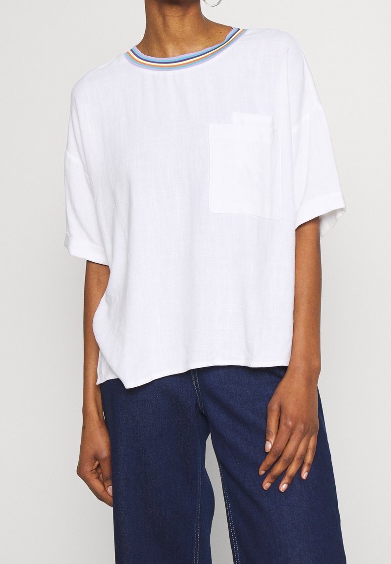 Personne portant un t-shirt blanc oversize avec une poche poitrine et un col rayé arc-en-ciel, associé à un jean bleu foncé.