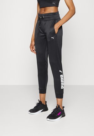 Mujer con pantalones deportivos negros Puma con logo blanco en la pierna y zapatillas negras con suelas blancas y logo morado, de pie frente a un fondo liso.