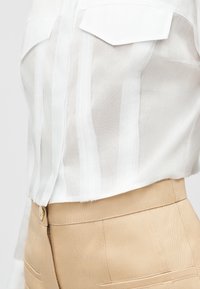 Λευκό διαφανές πουκάμισο με κάθετες ρίγες, μπερδεμένο σε μπεζ ψηλόμεσο παντελόνι με μπροστινές τσέπες και κλείσιμο με κουμπιά.