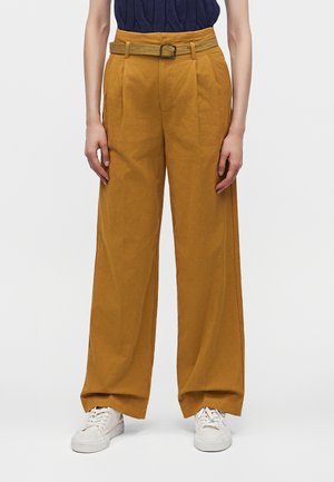 Persona que lleva pantalones anchos de color mostaza con un cinturón a juego y zapatillas blancas, de pie con los brazos relajados a los lados.