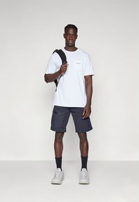 Valkoinen lyhythihainen t-paita, jossa on etutasku, yhdistettynä tummiin cargohousuihin, joissa on vetoketjutaskut, ja harmaat lenkkarit. Reppu olalla.