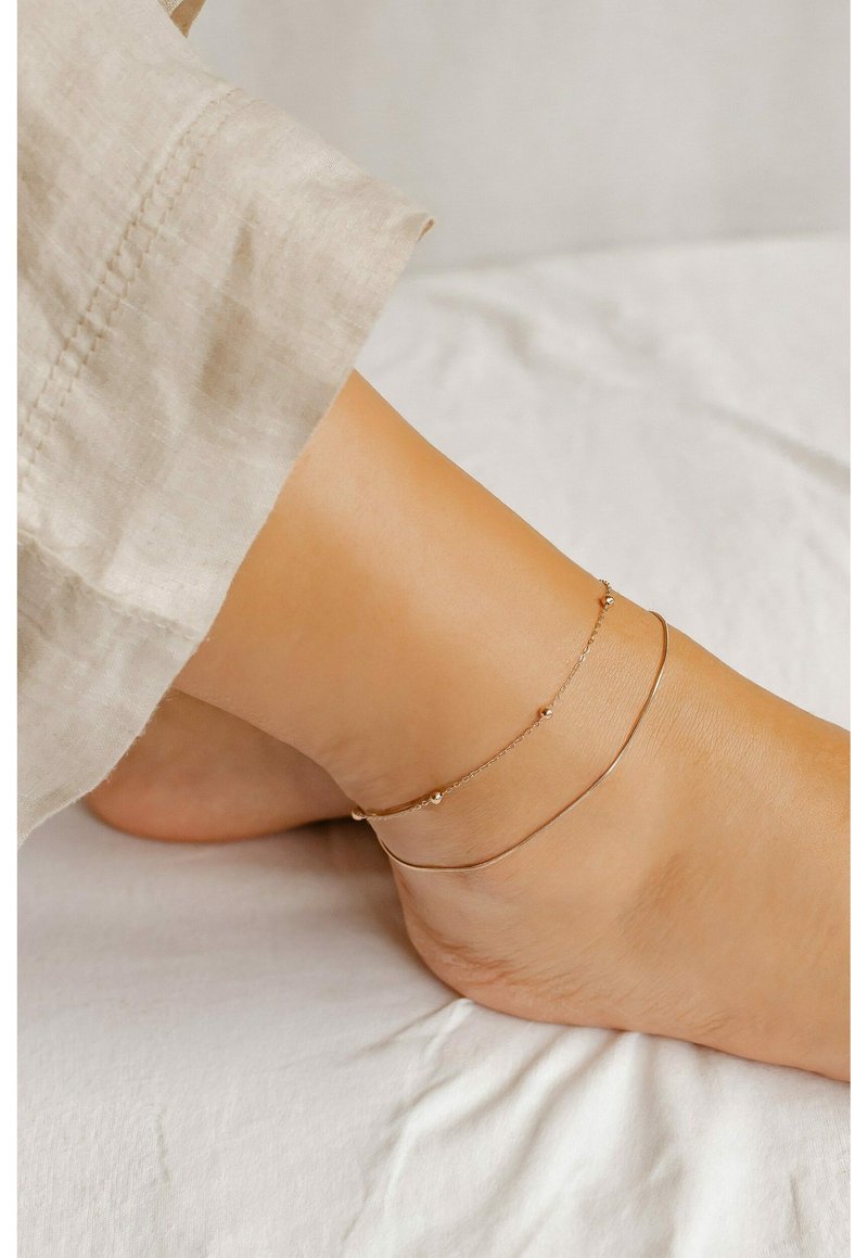 Cavigliera in tonalità oro con un delicato design a catena, caratterizzata da piccoli accenti circolari. Indossata su un piede nudo contro uno sfondo di tessuto chiaro.