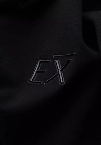 Sort stof med en glat tekstur og et blankt, sort logo "EX" prægede i midten. Simpelt og moderne design.