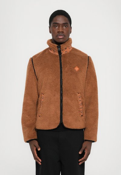 Junger Mann trägt eine braune Fleece-Jacke mit Reißverschluss und gemustertem Innenkragen sowie schwarze Hosen, steht vor einem hellen Hintergrund.