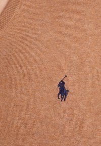 Pull en maille à col en V couleur beige chaud avec une texture douce. Présente un petit logo brodé en bleu marine représentant un joueur de polo sur le côté gauche de la poitrine.