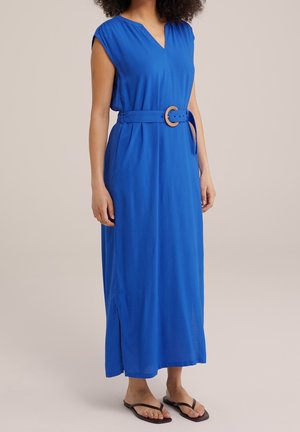 Femme portant une robe longue bleu sans manches avec une ceinture à la taille et des tongs noires, debout devant un fond neutre.