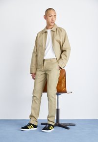Beige bomberjakke over en hvit t-skjorte, kombinert med beige bukser og svarte sneakers med gule striper. Enkel design og rene linjer.