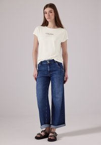 Lyscreme t-shirt med teksten "EVERYTHING", parret med brede blå jeans med opsmøg og sorte sandaler med sølvdetaljer.