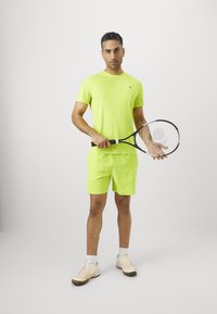 Mees, kes kannab erksat roheline spordivormi ja hoiab tennisereketit mõlemas käes, seisab ühtlase valge tausta ees.