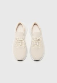 Baskets en maille beige avec une tige texturée, des lacets élastiques et une semelle blanche rembourrée. Conçues pour un ajustement confortable avec une esthétique minimaliste.