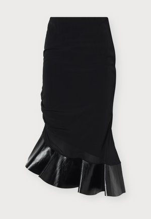Mujer de pie con un vestido negro de manga corta, detalle fruncido en la cintura y bajo brillante y acampanado, combinado con zapatos negros puntiagudos.