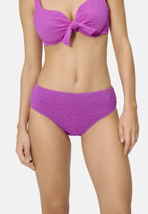 Mujer vistiendo un bikini brillante de color púrpura texturizado, con un lazo frontal en la parte superior y braguitas de cintura alta, contra un fondo claro y sencillo.