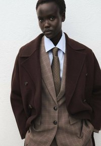 Un manteau en laine marron superposé sur un costume à carreaux avec une chemise habillée bleue et une cravate à motifs. L'ensemble présente des détails sur mesure et des boutons.