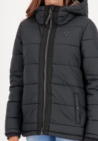 Vrouw in een zwarte, gewatteerde jas met capuchon, voorzien van een rits aan de voorkant, drukknopen zakken en een klein hart-embleem op de borst.