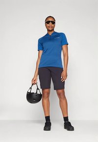 Modra kolesarska majica s kratkimi rokavi in zadrgo do polovice, kombinirana s črnimi hlačami in črnimi čevlji. Model drži črno čelado v eni roki.