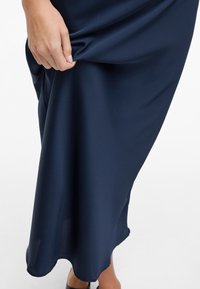 Kraljevsko modra satenasta obleka z gladko teksturo, ki ima tekoč dizajn in valovit rob, ki jo rahlo drži roka blizu pasu.