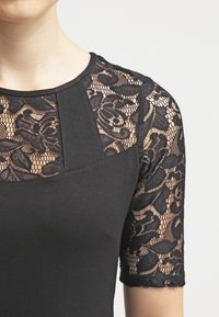 Svart topp med en figurnära passform och korta ärmar, dekorerad med intrikat svart spets. Spetsen har blommönster och en slät tyg nedanför.