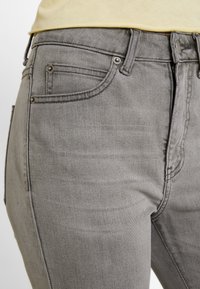 Jeans de mezclilla gris con un diseño clásico de cinco bolsillos, con herrajes metálicos y una textura sutil, mostrados de lado.