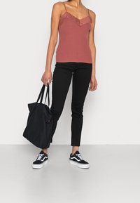 Camisola rosa con pizzo, jeans neri aderenti e borsa a tracolla nera. Modella che indossa sneaker bianche e nere, in piedi su uno sfondo bianco.