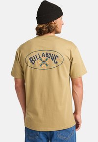 Beige katoenen t-shirt met een groot gedrukt logo in zwart en geel op de rug, met een ronde omtrek en decoratieve elementen.