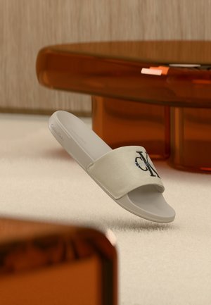 Fehér Calvin Klein papucs fekete logóval a szövetpánton, világos szőnyegen, áttetsző barna bútor mellett elhelyezve.