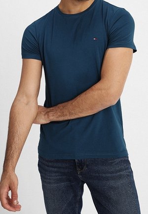 Person trägt ein eng anliegendes dunkelblaues T-Shirt mit kleinem Logo auf der Brust und blaue Jeans, die linke Hand berührt den rechten Unterarm.