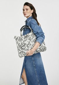 Duża torba na ramię z kwiatowym wzorem w czerni i bieli, wykonana z materiału, posiada podwójne czarne uchwyty oraz sztywną formę, trzymana przez modela.