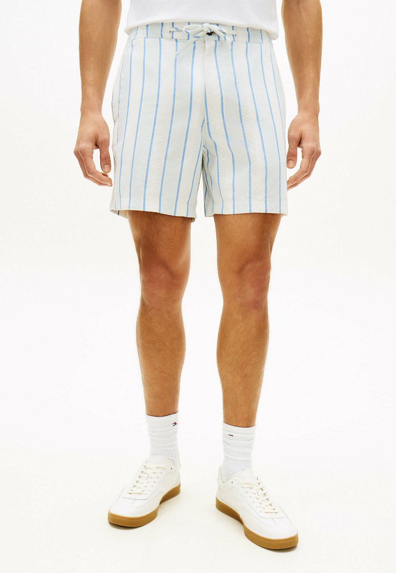 Uomo con pantaloncini bianchi a righe verticali blu, calzini bianchi alla caviglia e scarpe da ginnastica bianche in piedi su uno sfondo bianco.